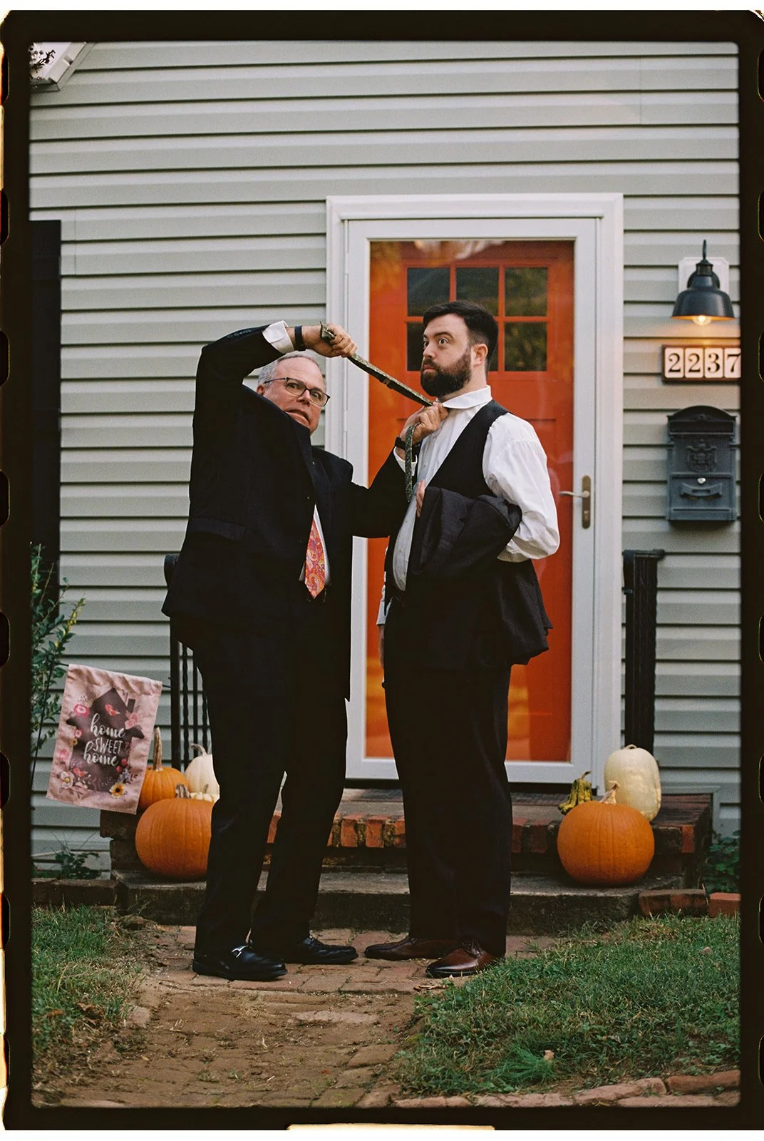 A man with glasses and a man with a beard stand in front of a house with a red door. The man with glasses is pulling a tie around the other man's neck. There are pumpkins and a bag with fall decorations on the porch. Roanoke Virginia Wedding Photo