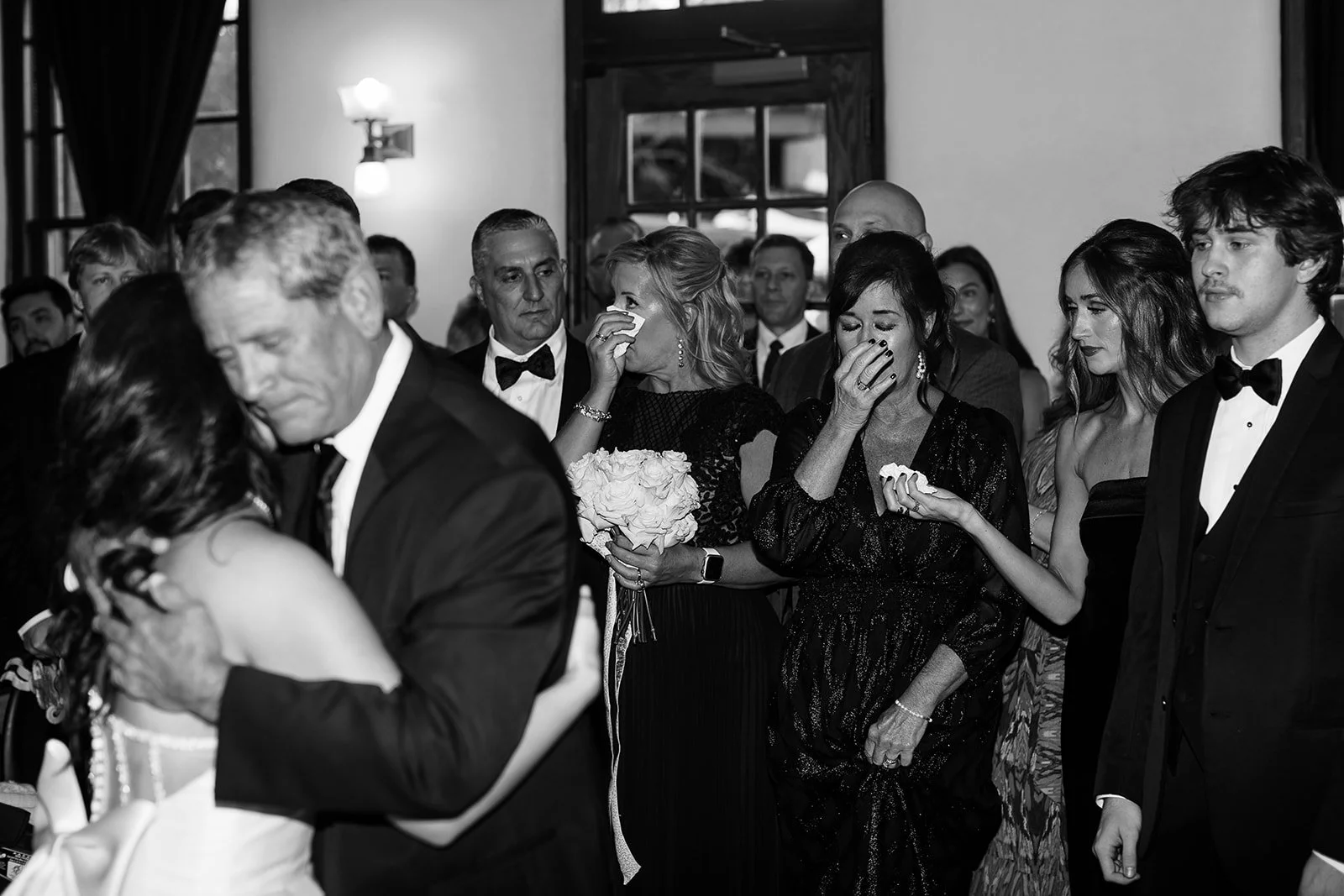 Black and white photo of emotionally overwhelmed wedding guests, mostly women, wiping tears, holding tissues and flowers, attending a couple's wedding. roanoke wedding film photographer virginia