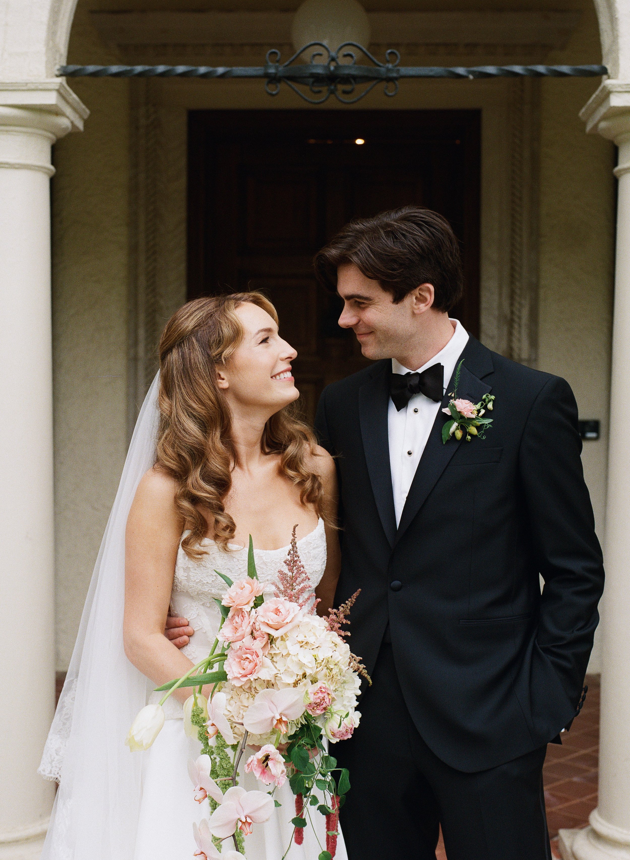 A bride and groom standing close together, smiling and looking into each other's eyes, in front of a building with columns and an arched entrance, for a wedding ceremony.