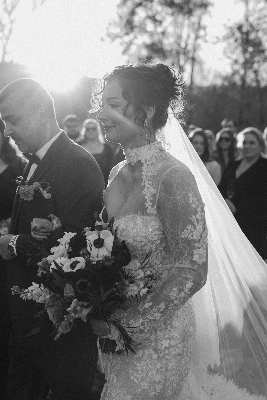 A black and white photo of a bride in a lace wedding dress holding a bouquet, standing outdoors during a wedding ceremony, with a groom beside her and guests in the background. potters craft cider charlottesville va