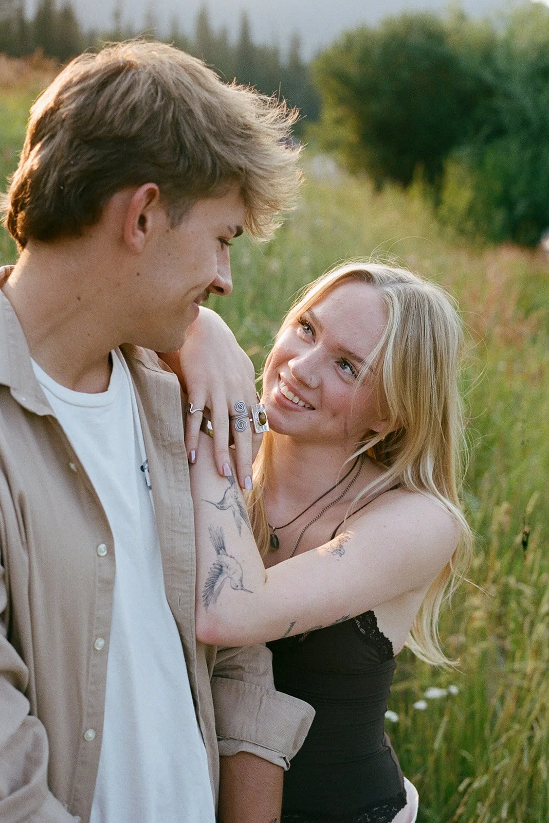 A young couple smiling and looking at each other outdoors in a field with greenery. bozeman engagement photographer
