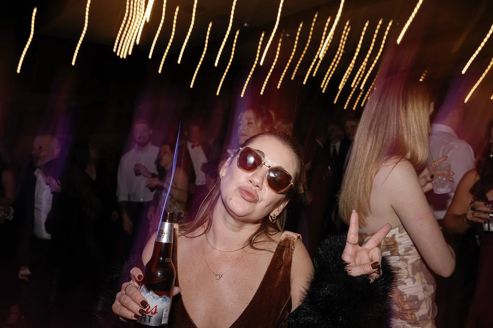 A woman wearing sunglasses and holding a beer at a party or nightclub with colorful lights and other people in the background. wintergreen resort Wedding Film Photographer