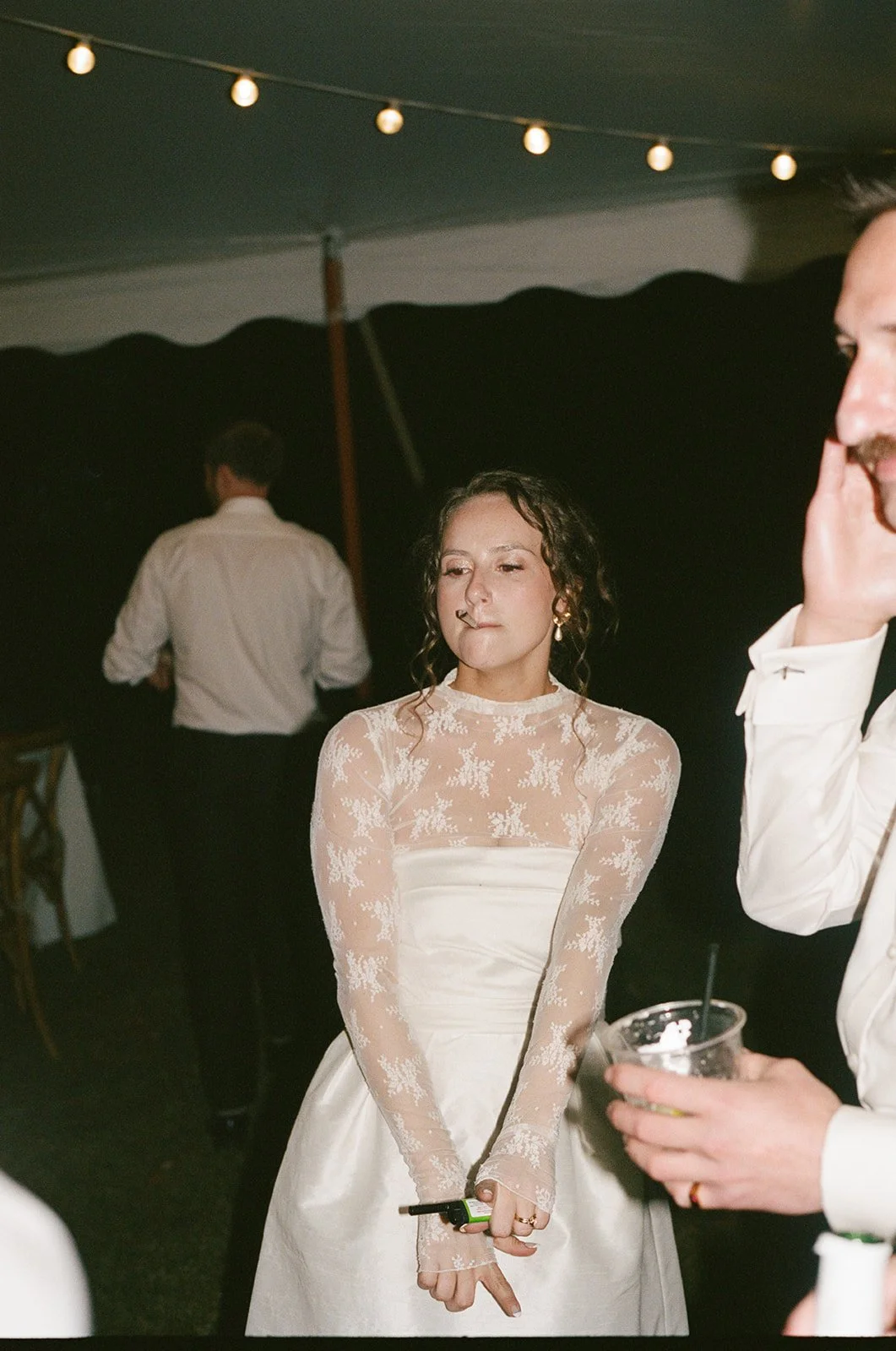 A woman in a white lace long-sleeve dress with dark curly hair standing at a social event, holding a lighter in her right hand, with string lights hanging overhead. Charlottes Virginia Wedding Film Photographer