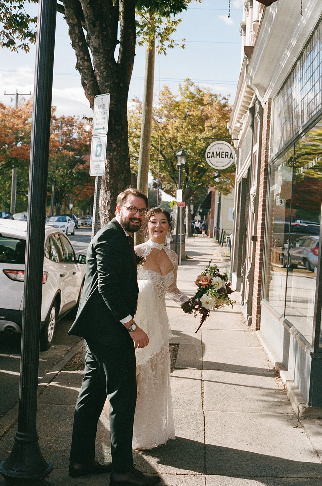 A bride and groom standing on a sidewalk in front of a photography studio, smiling and looking at the camera, with trees and parked cars visible in the background. downtown charlottesville virginia