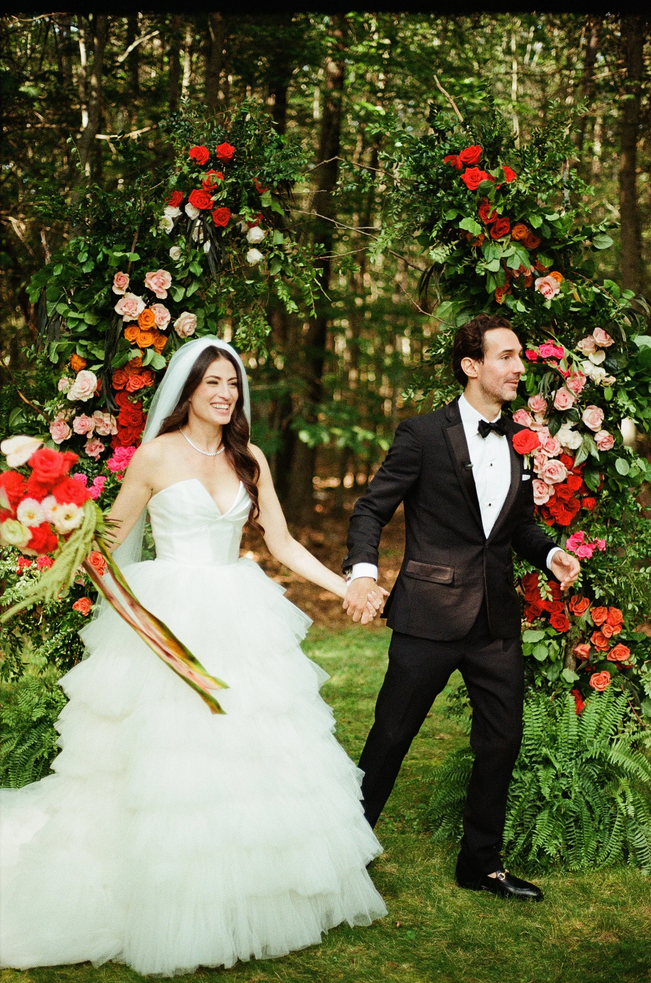 Bride and groom walking through an outdoor floral arch, surrounded by colorful flowers and greenery, in a wooded area in Portland Maine. The bride is in a white wedding gown holding a bouquet, and the groom is in a black tuxedo.