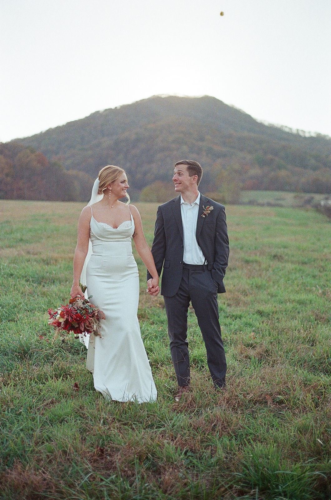A bride and groom holding hands and walking in a grassy field with a mountain in the background, during their wedding. roanoke wedding film photographer