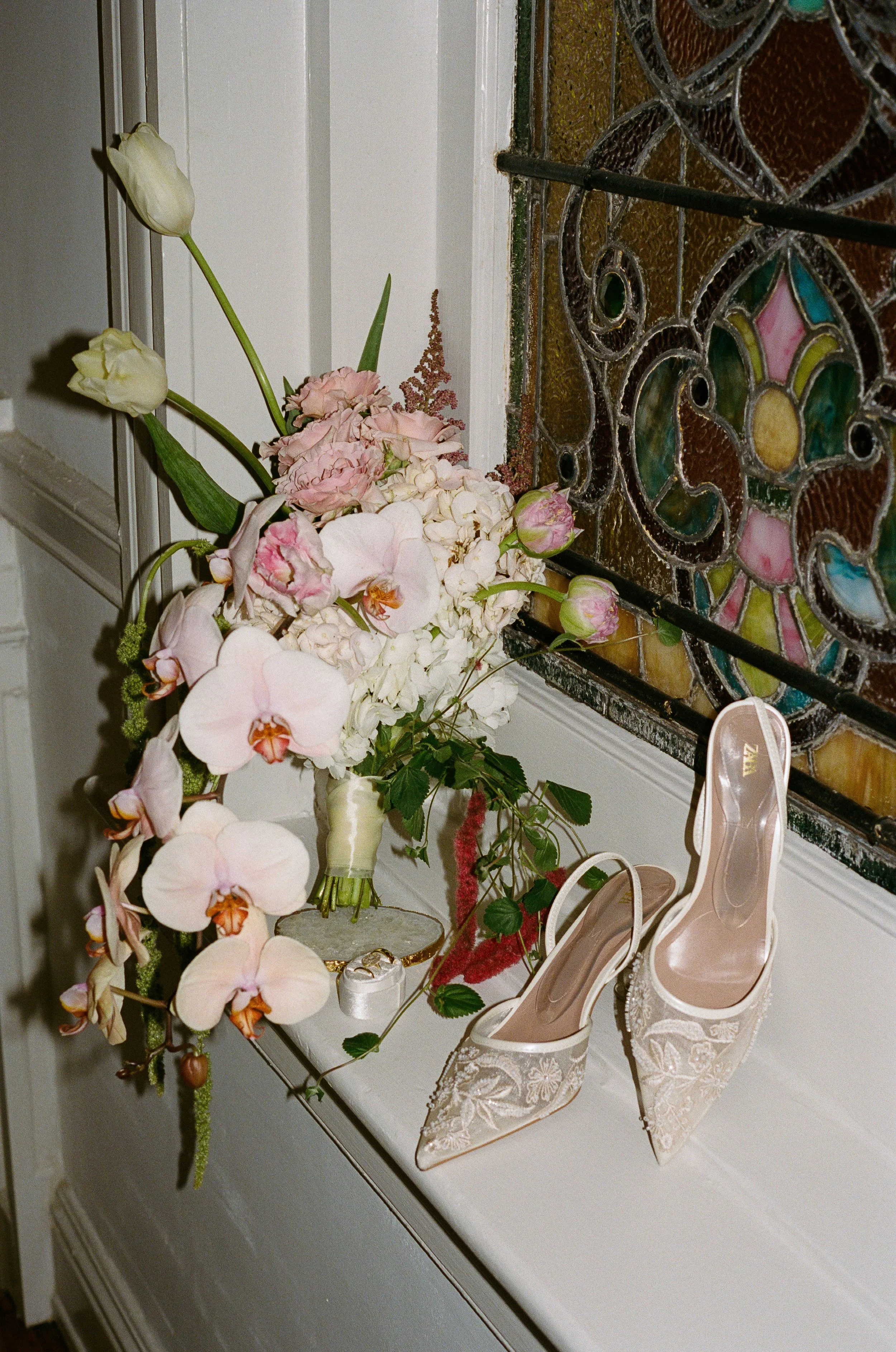 A floral arrangement with pink orchids and white flowers placed on a table near a stained glass window, with a pair of transparent high-heeled shoes with white lace details on the floor. richmond virginia wedding photographer