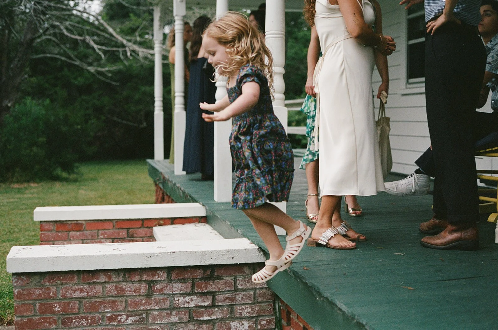 A young girl jumps off a small porch onto the grass, with a group of adults standing nearby on a porch. ocracoke outerbanks wedding film photographer