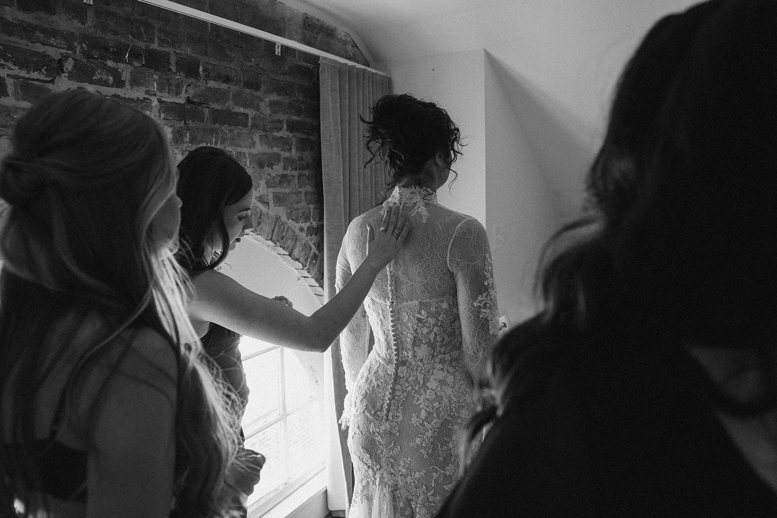 A bride in a lace wedding dress standing by a window as friends help her prepare, with one friend touching her shoulder, in a black-and-white photo inside a room with an exposed brick wall. doyle hotel charlottesville va