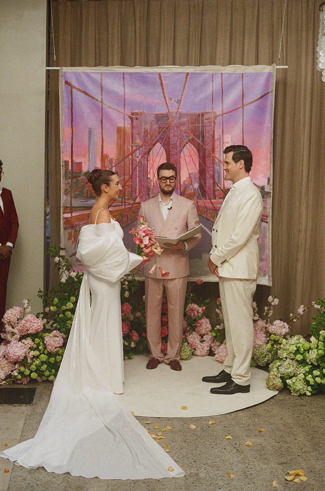 A wedding ceremony with a bride in a white gown holding pink flowers, and a groom in a cream suit, standing in front of an officiant in a pink suit. Behind them, a backdrop of a painting depicting a bridge, with floral arrangements surrounding the scene.