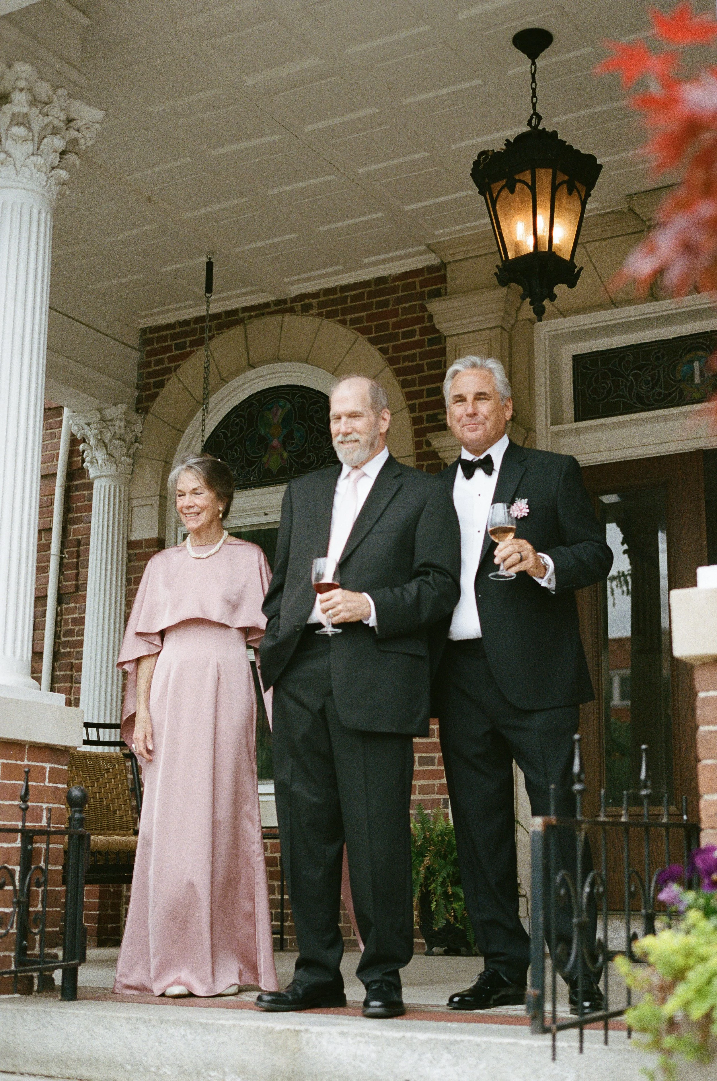Three well-dressed people, two men in tuxedos and a woman in a pink gown, standing outdoors on the porch of a brick house with white columns and stained glass windows, holding wine glasses.
