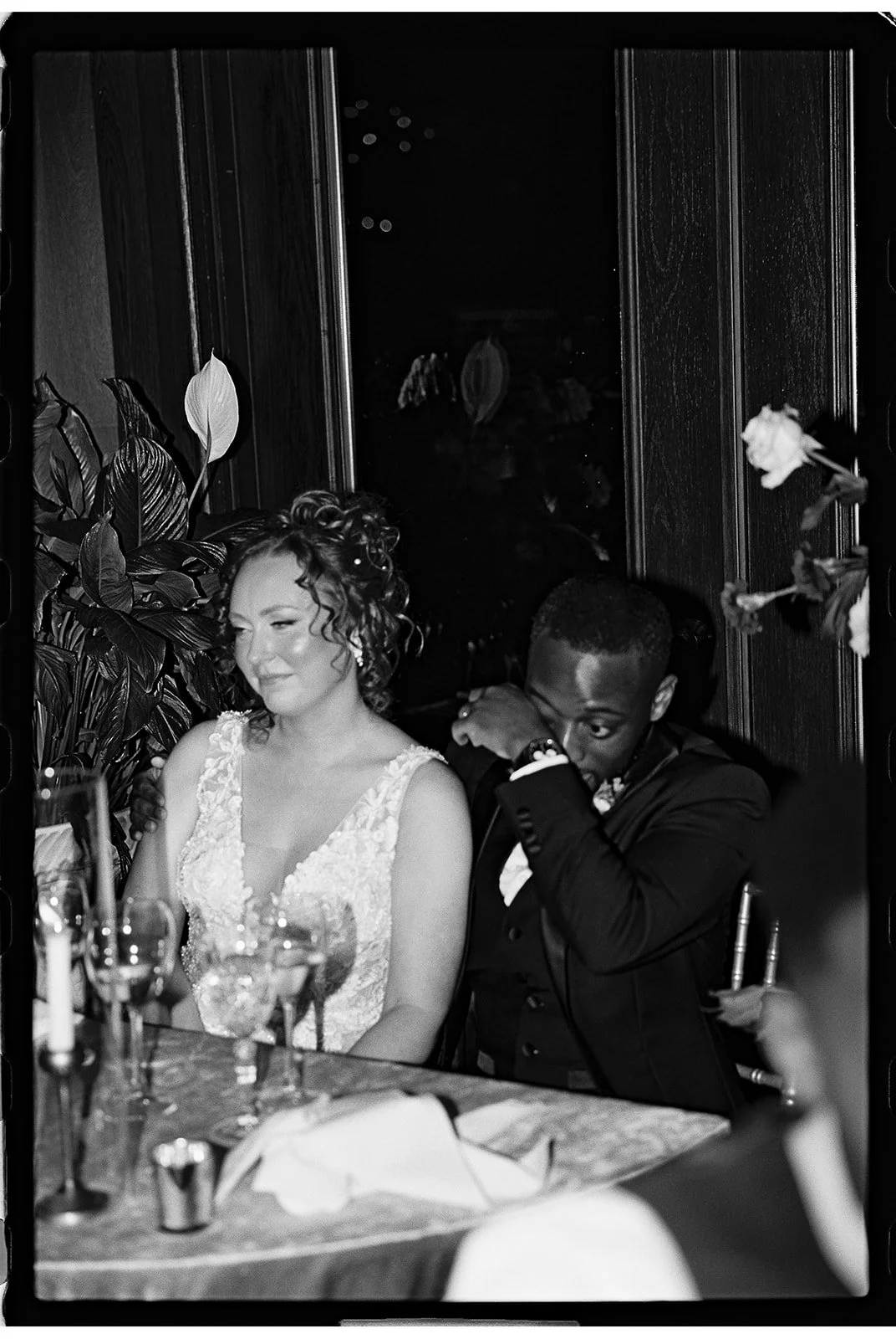 A woman in a wedding dress and a man in a tuxedo sitting at a dinner table during a formal event or wedding reception. The woman is smiling and the man has his head bowed and appears to be wiping his eye. There are glasses and a napkin on the table, 