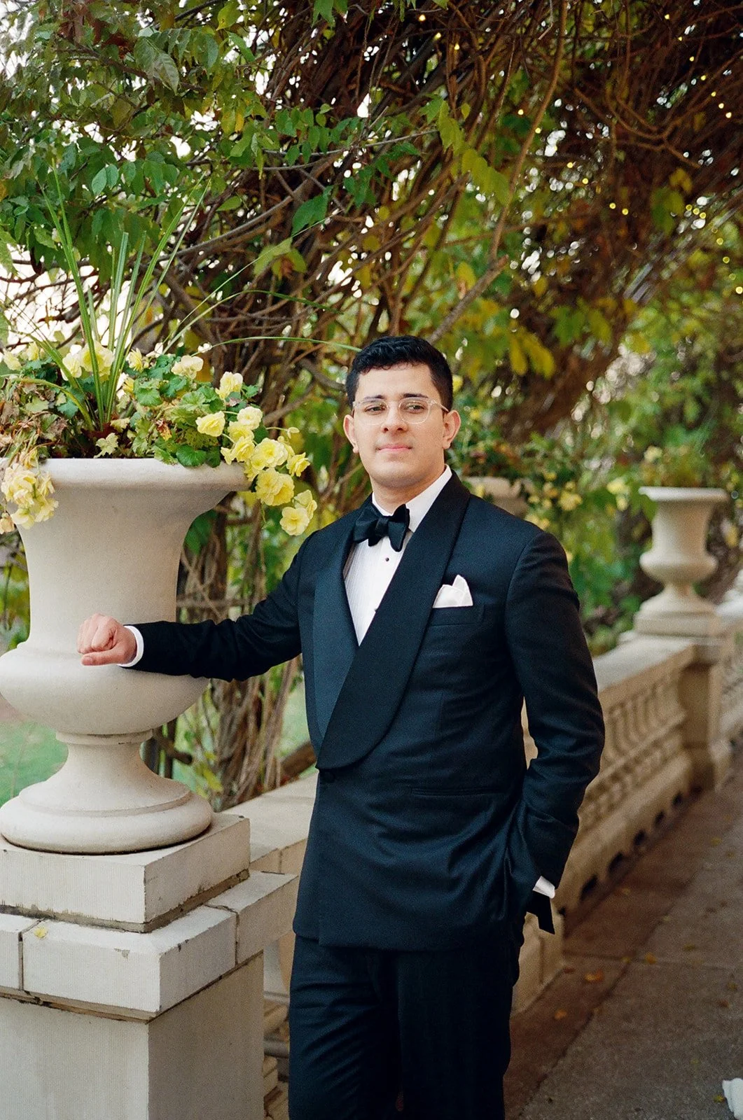 A young man in a black tuxedo with a white dress shirt and black bow tie, standing outdoors beside a stone railing and an ornate large white planter filled with yellow flowers, with leafy greenery in the background. liriodendron mansion
