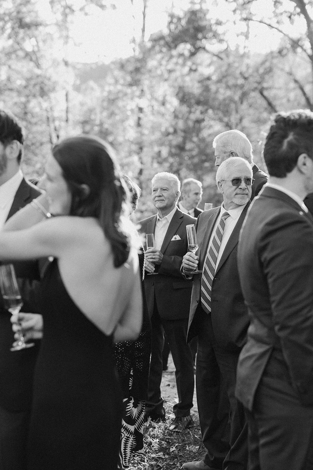 A black and white photo of people outdoors at a gathering, holding champagne glasses, dressed in formal attire, with trees and sunlight in the background. potters craft cider charlottesville va