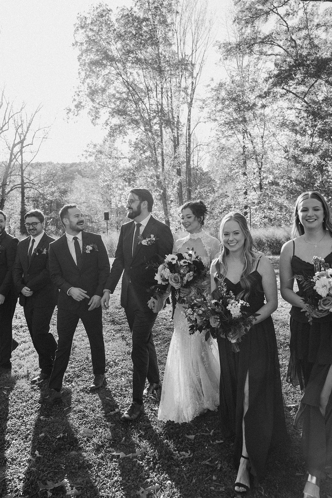 A black and white photo of a wedding party outdoors with tall trees in the background, featuring the bride and groom walking arm-in-arm surrounded by bridesmaids and groomsmen, all smiling and holding bouquets. potters craft cider charlottesville va