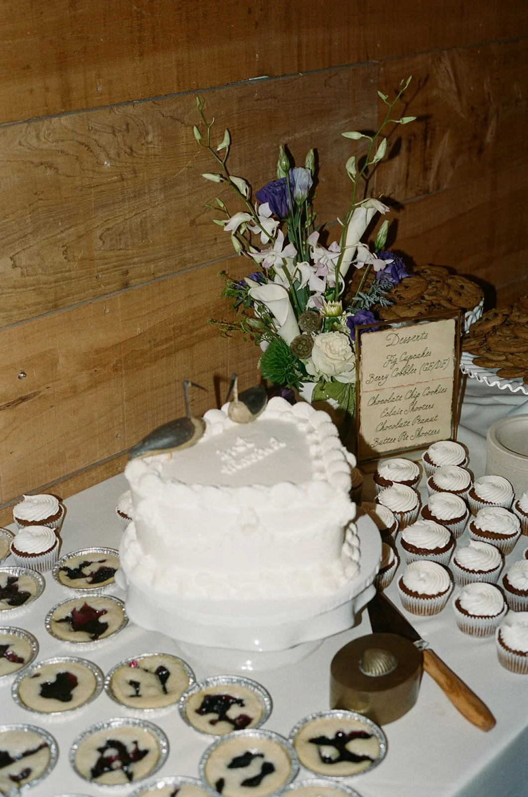 A dessert table with a wedding cake, cupcakes, and tarts, decorated with flowers and a wooden background. ocracoke outerbanks wedding film photographer