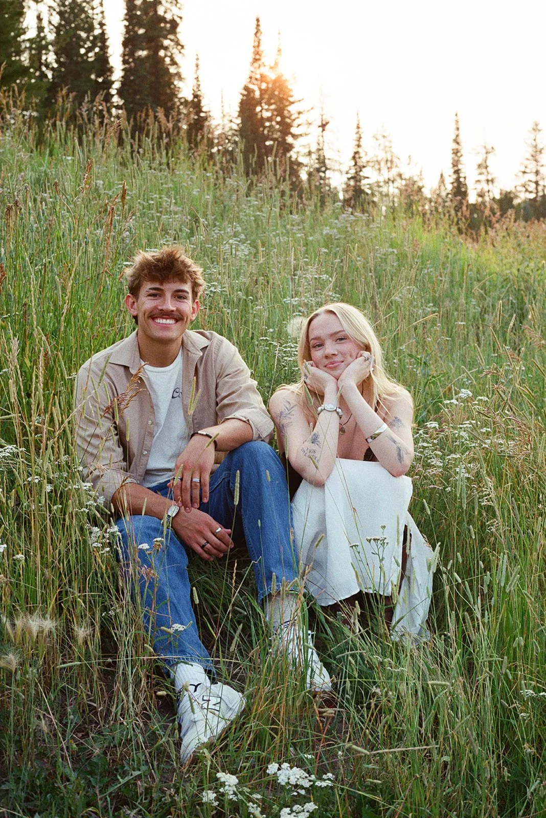 A young man and woman sitting in a grassy field with wildflowers, smiling at the camera, during sunset. bozeman montana