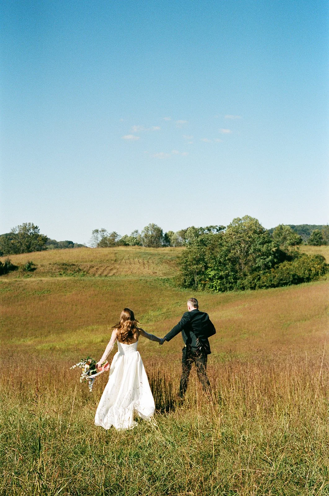 A bride and groom holding hands and walking through a grassy field under a clear blue sky, with trees and rolling hills in the background. roanoke wedding film photographer
