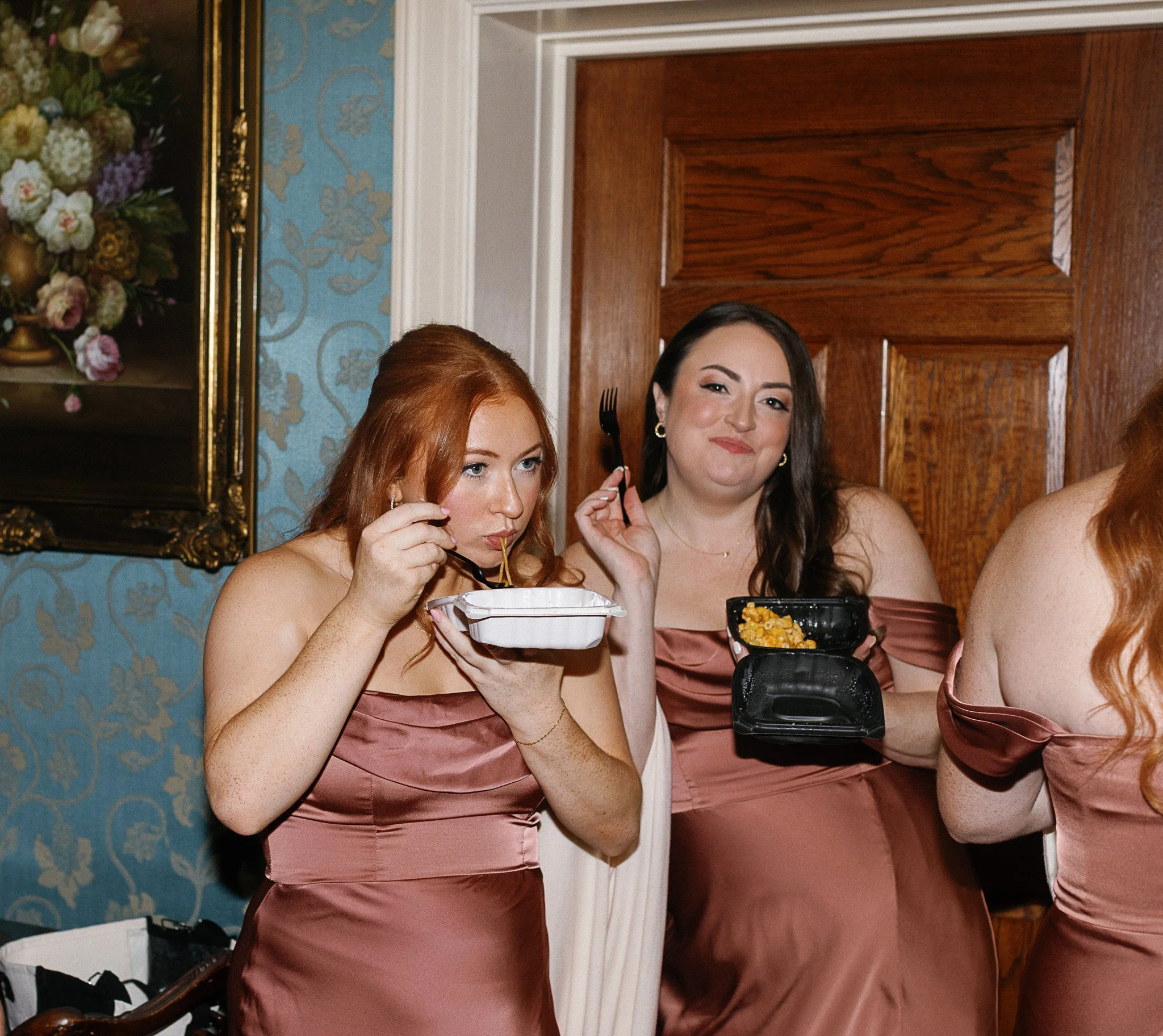 Two women in formal dresses eating food from takeout containers inside a room with floral wallpaper and a painting on the wall. gramercy mansion maryland wedding