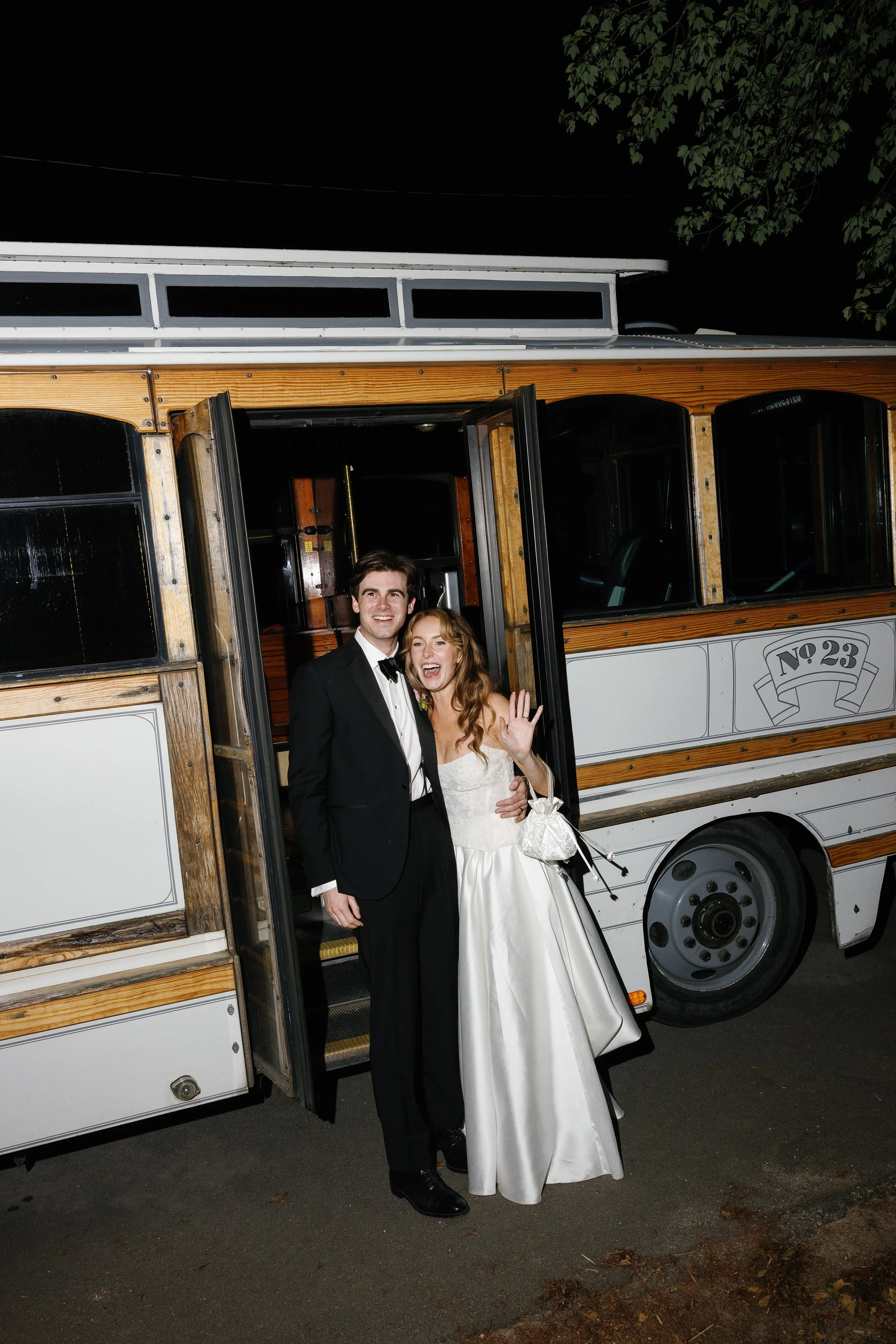 A man and woman in wedding attire standing in the doorway of a vintage trolley bus, smiling and waving at the camera at night.