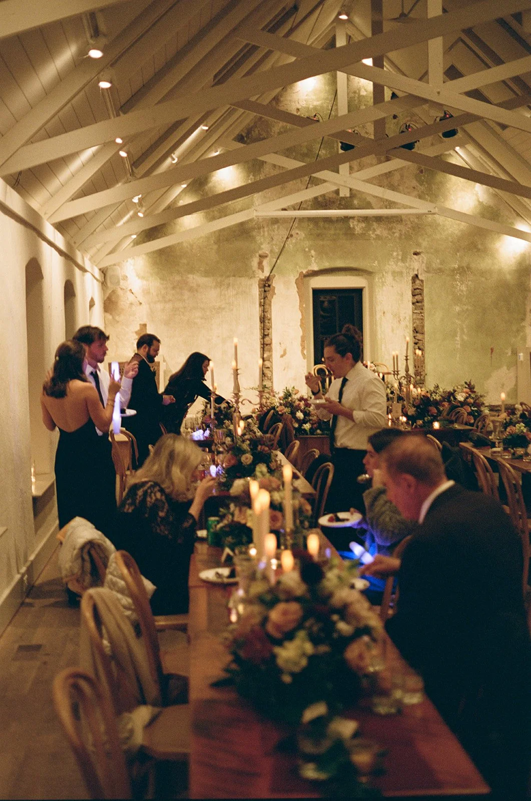 People dining at a long, elegant table with floral centerpieces and candlelight in a rustic, warmly lit room with exposed beams and distressed walls potters craft cider charlottesville va