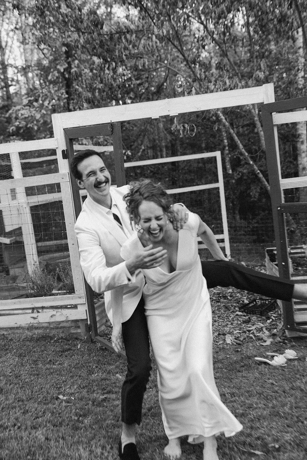 A joyful black and white photo of a man in a white suit and a woman in a white dress laughing and playing outdoors. Charlottesville Virginia Wedding Film Photographer