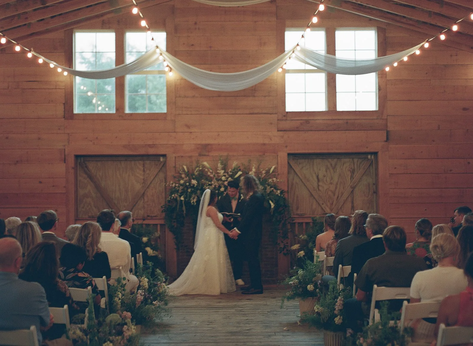 A wedding ceremony inside a rustic wooden barn. The couple stands at the altar, holding hands, facing each other, with the officiant reading from a book. The bride wears a long white dress with a veil, and the groom wears a dark suit. Guests are seat