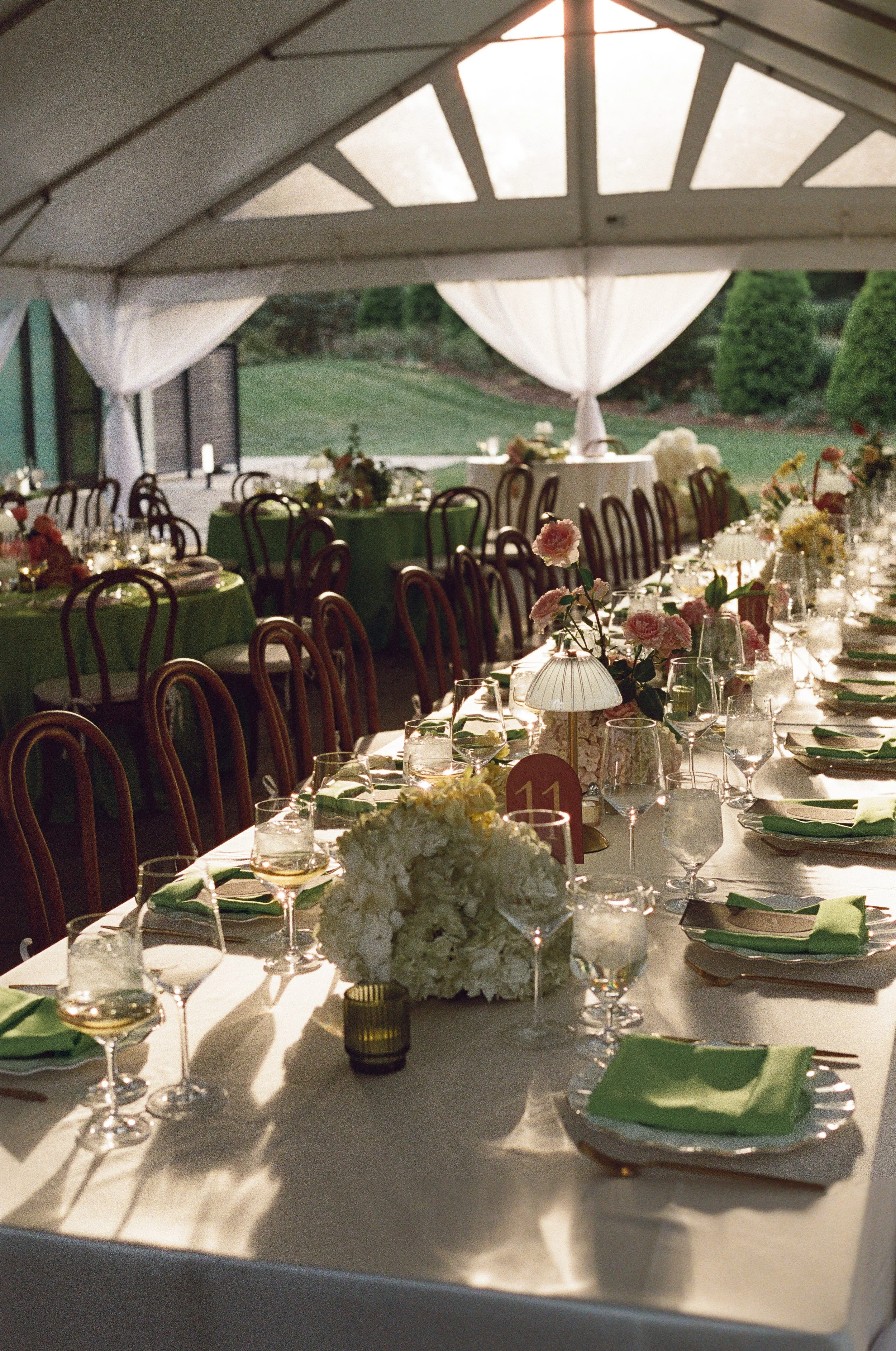 A decorated outdoor tent with tables set for a formal event, featuring floral centerpieces, tableware, and green napkins, with a view of a grassy landscape through the open sides.