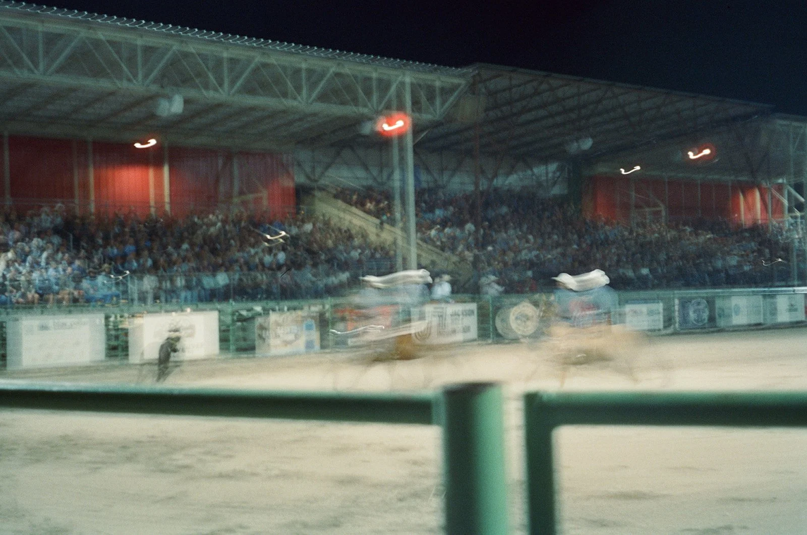 Nighttime rodeo with blurred horses and cowboys, grandstand filled with spectators, and a red-lit structure in the background. in Jackson Wyoming Film Photographer