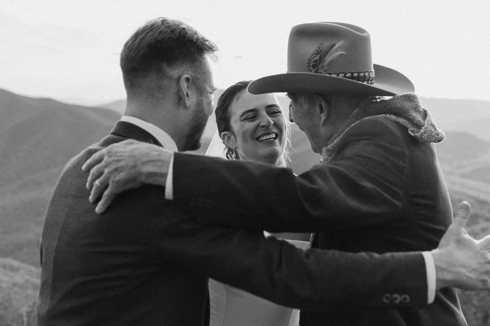 A joyful woman in a wedding dress smiling as she embraces two men, one in a suit and the other in a cowboy hat, outdoors with rolling hills in the background. wintergreen resort Wedding Film Photographer