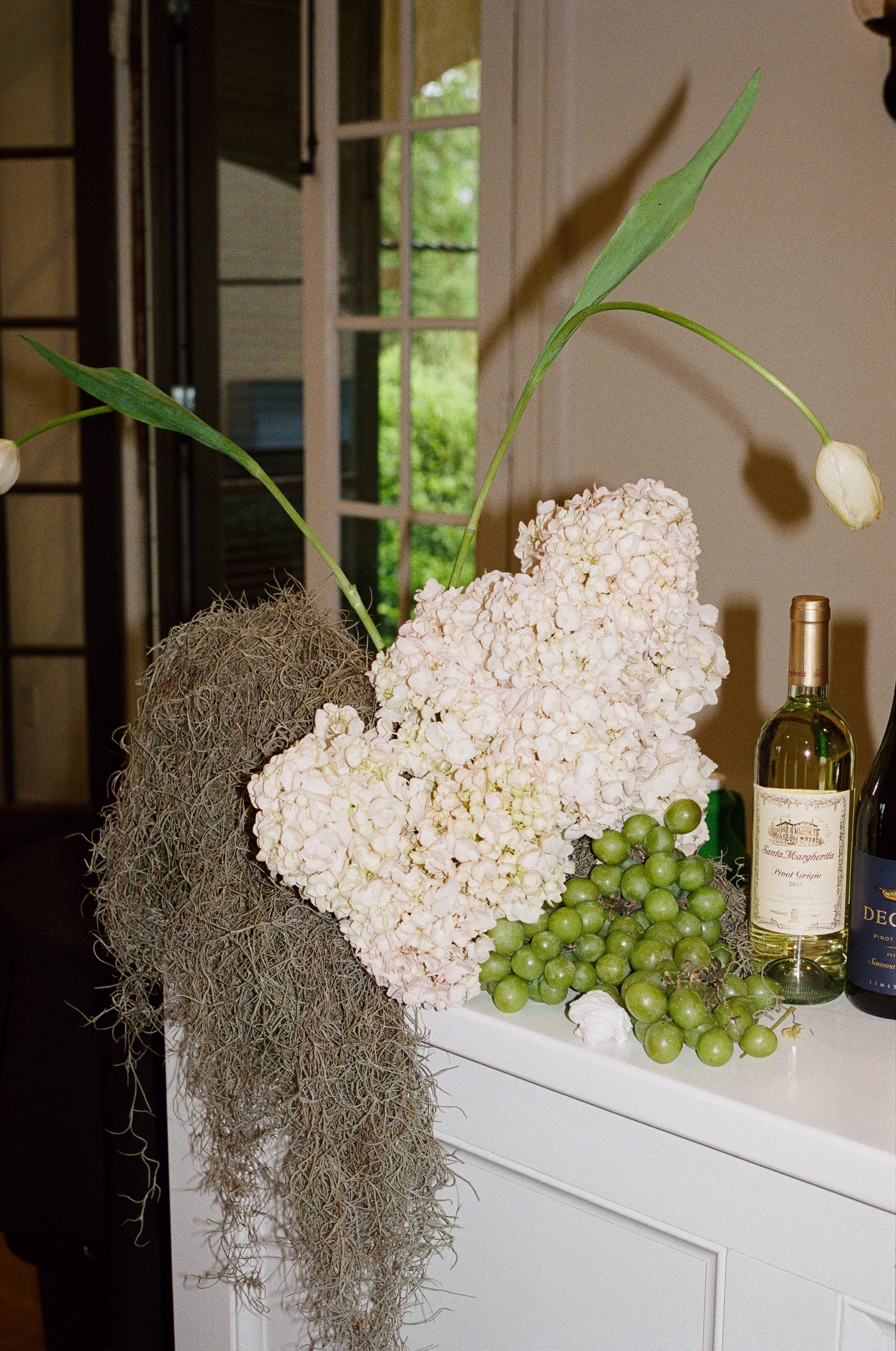 Arrangement of pink hydrangea flowers, green grapes, and an orchid plant with white flowers and green leaves, near a bottle of white wine on a white surface, with a window and greenery outside in the background.