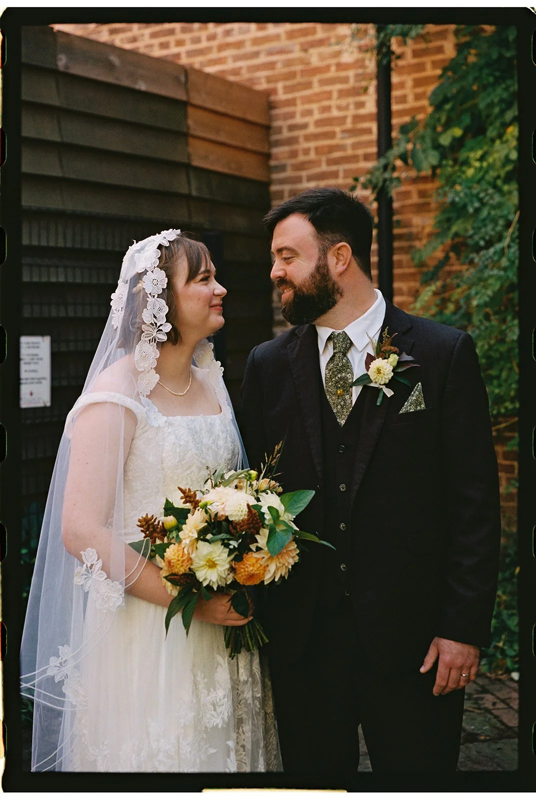 A bride and groom standing close together outdoors, smiling at each other. The bride is wearing a vintage lace wedding dress with a floral veil and holding a bouquet of yellow and white flowers. The groom is wearing a dark suit with a patterned tie, 
