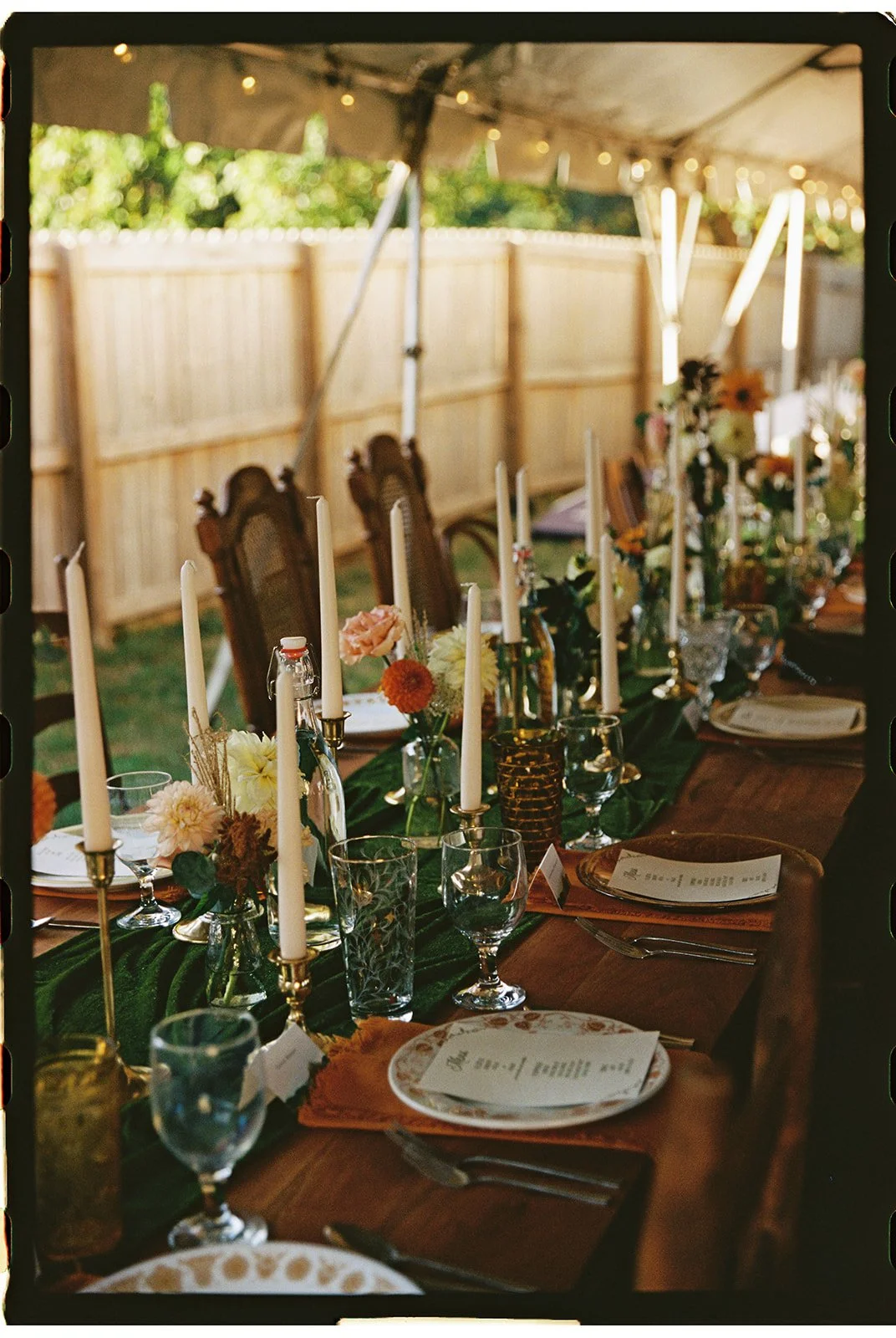 A decorated outdoor dining table with candles, flowers, glassware, and place settings, under a canopy with string lights and a wood fence in the background. Roanoke Virginia Wedding Film Photographer.