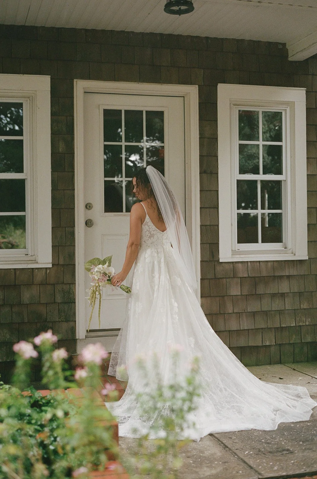 A bride in a white wedding gown holding a bouquet of flowers, standing on a porch with brown shingle siding, in front of a white door with glass panes and two windows on either side. ocracoke outerbanks wedding film photographer
