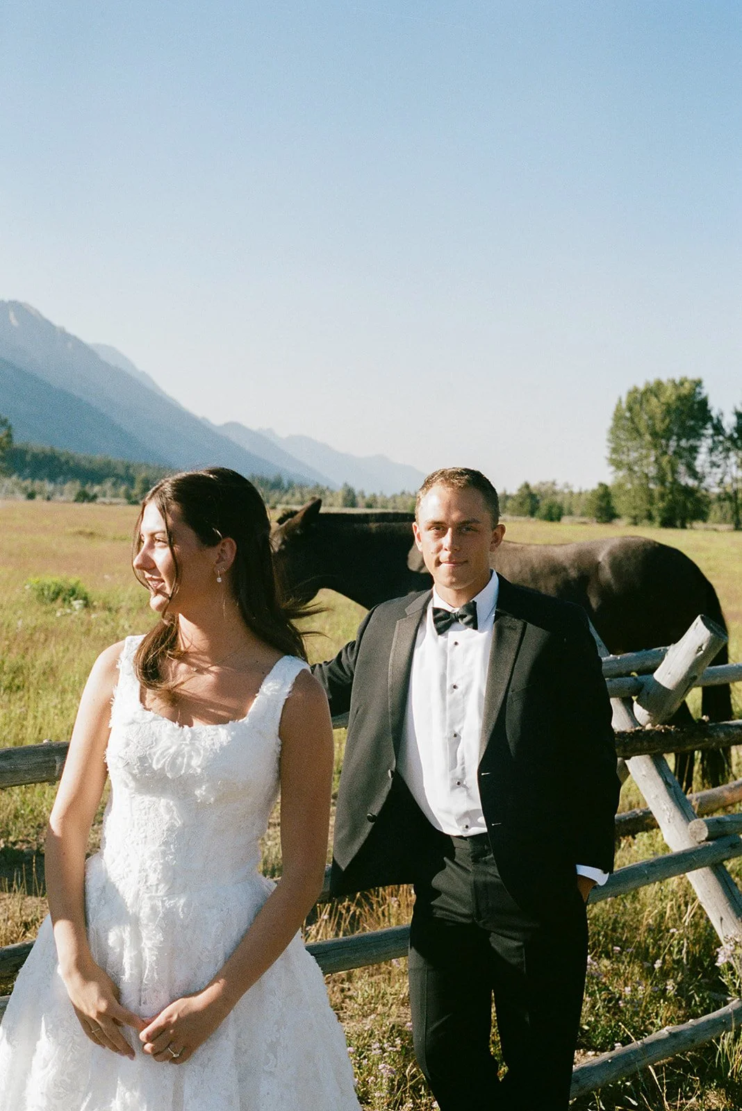 A bride and groom standing outdoors near a wooden fence, with a horse and mountains in the background on a sunny day. jackson hole wyoming wedding photographer