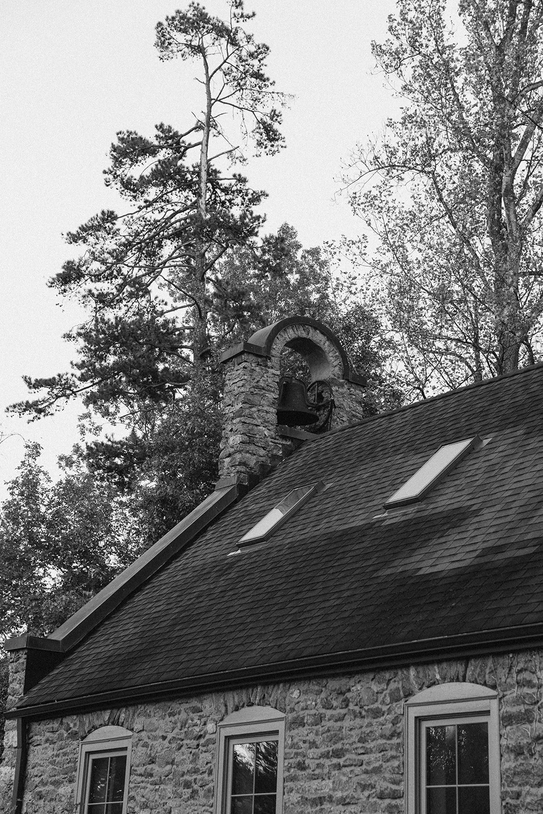 Black and white photograph of a stone house with a steeply sloped roof, two skylights, and a brick chimney with a bell and bell arch. Tall trees are visible in the background. potters craft cider charlottesville va