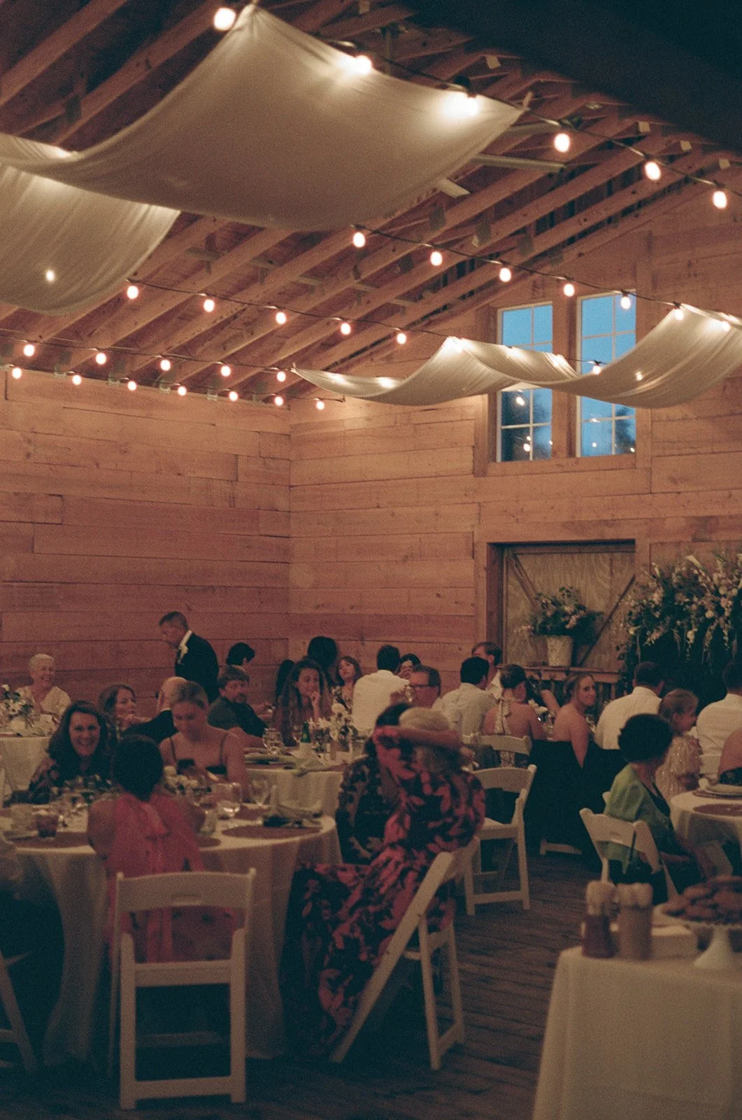 People gathered at a wedding reception in a rustic barn with wooden walls and string lights hanging from the ceiling. ocracoke outerbanks wedding film photographer