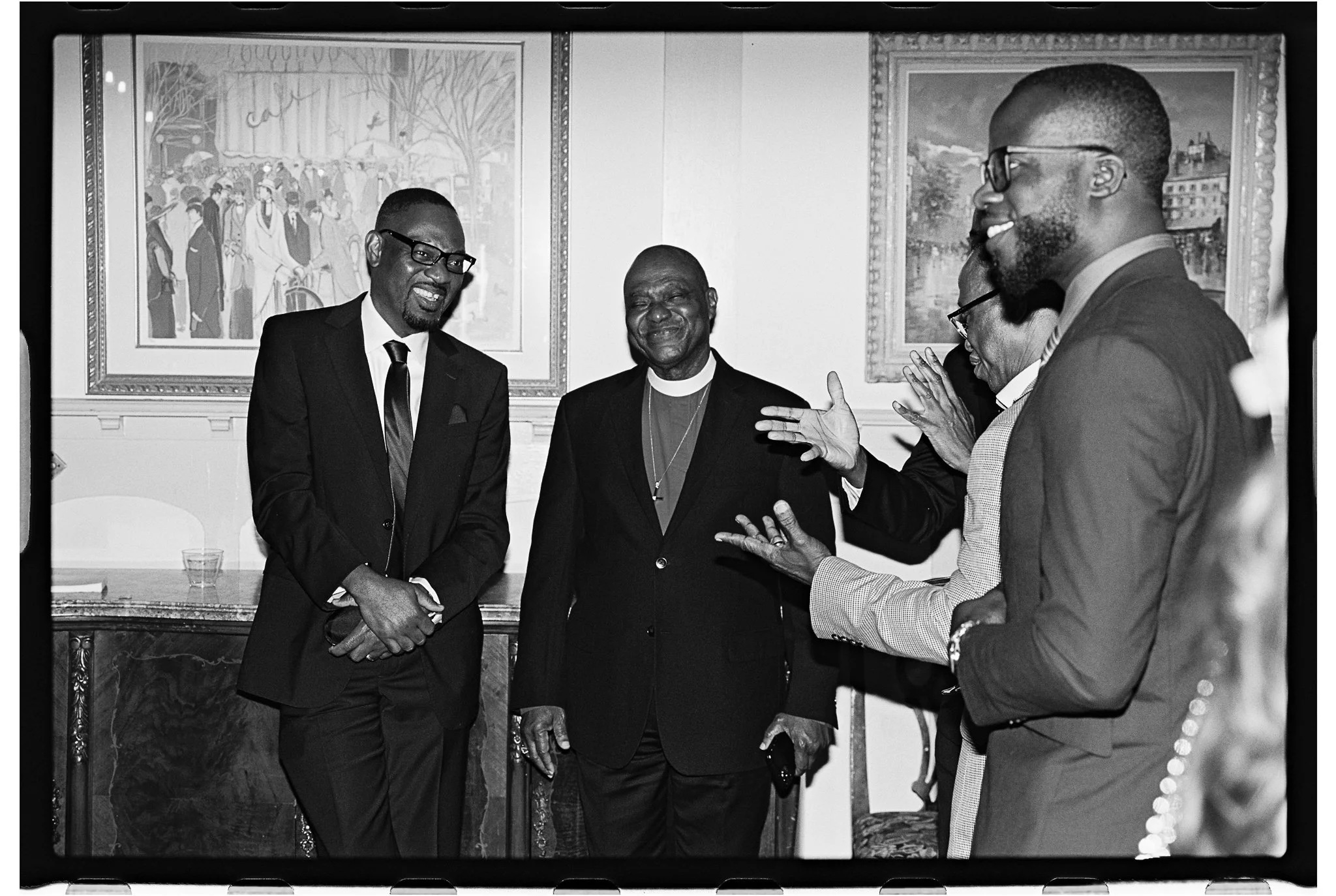 Black and white photo of a group of men in suits conversing and smiling indoors gramercy mansion maryland wedding 35mm film