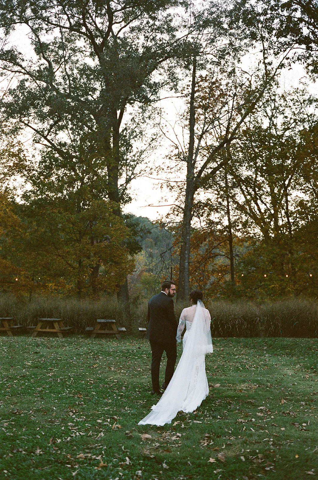 A couple dressed in wedding attire walking outdoors in a park surrounded by trees with autumn foliage. potters craft cider charlottesville va