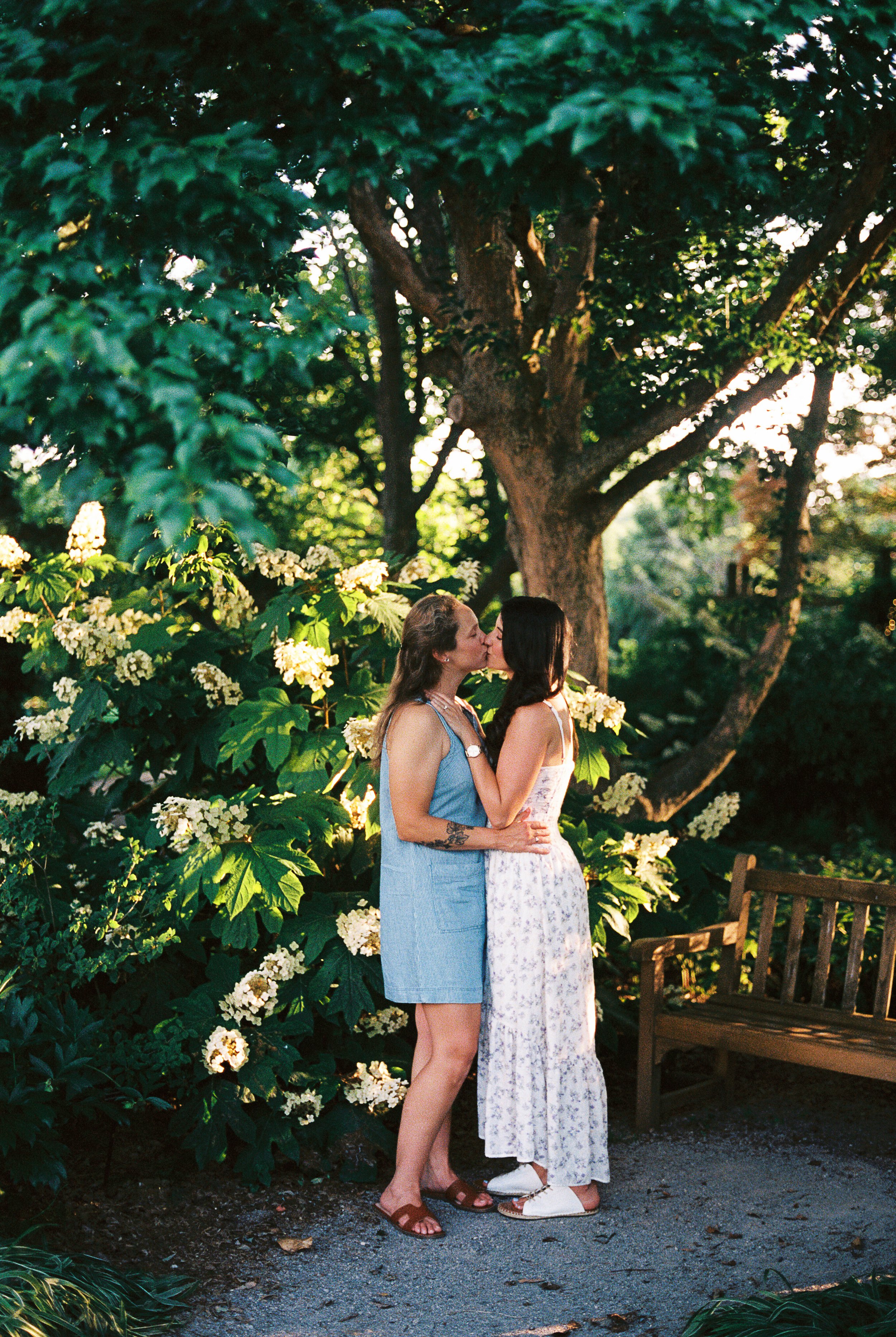 Two women kissing in a garden with green foliage, white flowers, and a wooden bench at Hahn Horticulture Garden, Virginia Tech. Blacksburg Virginia.