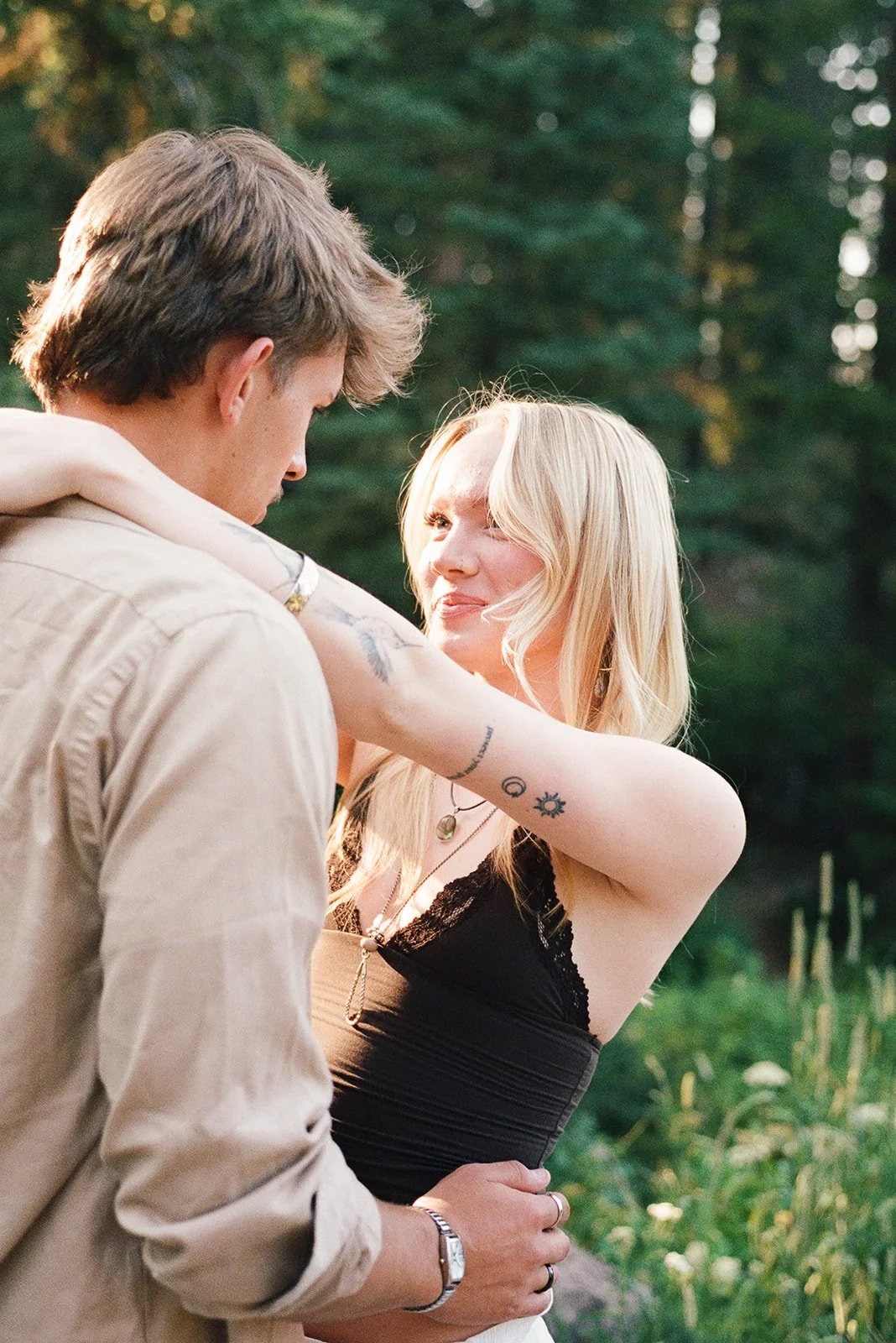 A couple standing outdoors in a forested area, with the man looking down and the woman smiling at him. The woman has long blonde hair and tattoos on her arm. bozeman montana wedding photographer