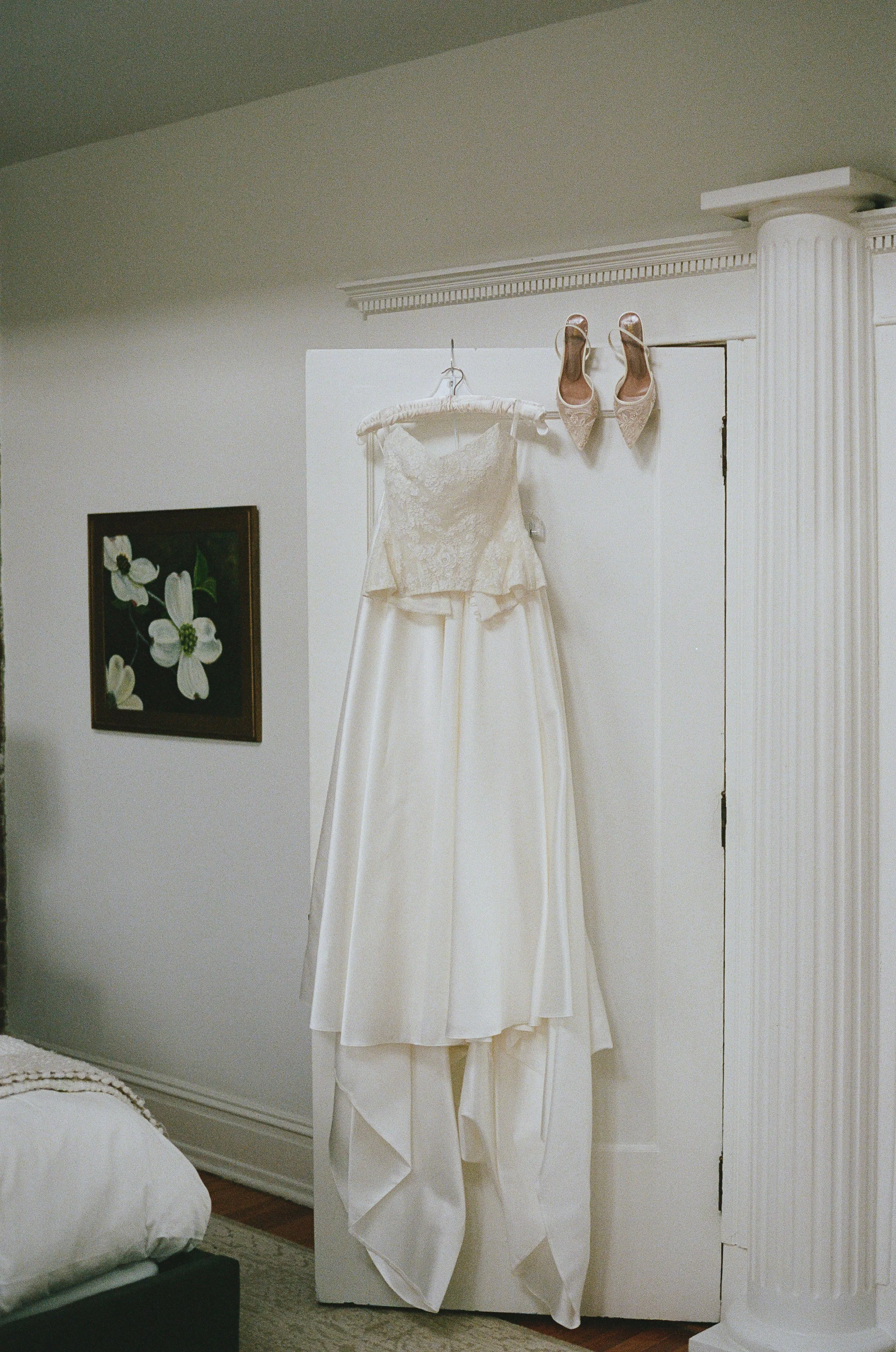 A wedding dress is hanging on a hanger on the corner of a white wall, with a pair of beige high-heeled shoes hanging on the wall above the dress, and a framed floral painting on the left side of the wall.