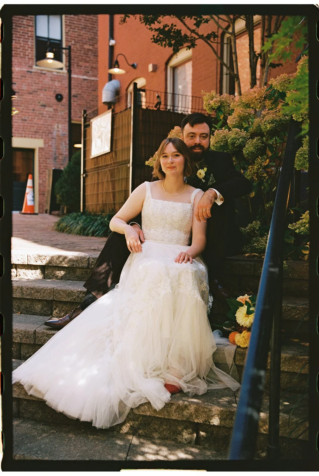 A bride and groom sitting on outdoor stone steps, with the bride in a white wedding dress and the groom in a black suit, surrounded by greenery and flowers. Staunton Virginia Wedding Film Photographer.