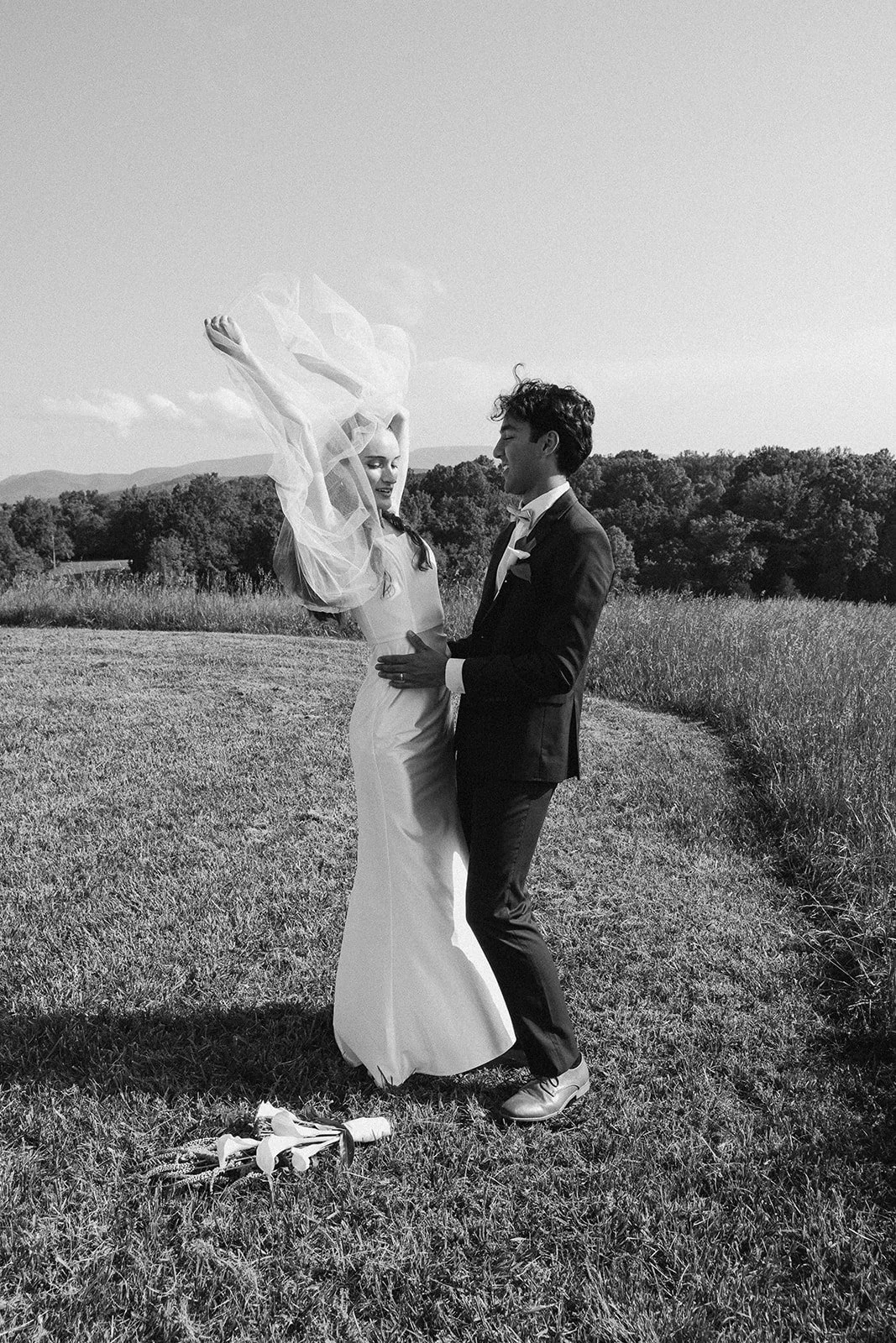 A black-and-white photo of a bride and groom dancing outdoors in a grassy field, with trees and hills in the background. Lexington Virginia Wedding Film Photographer