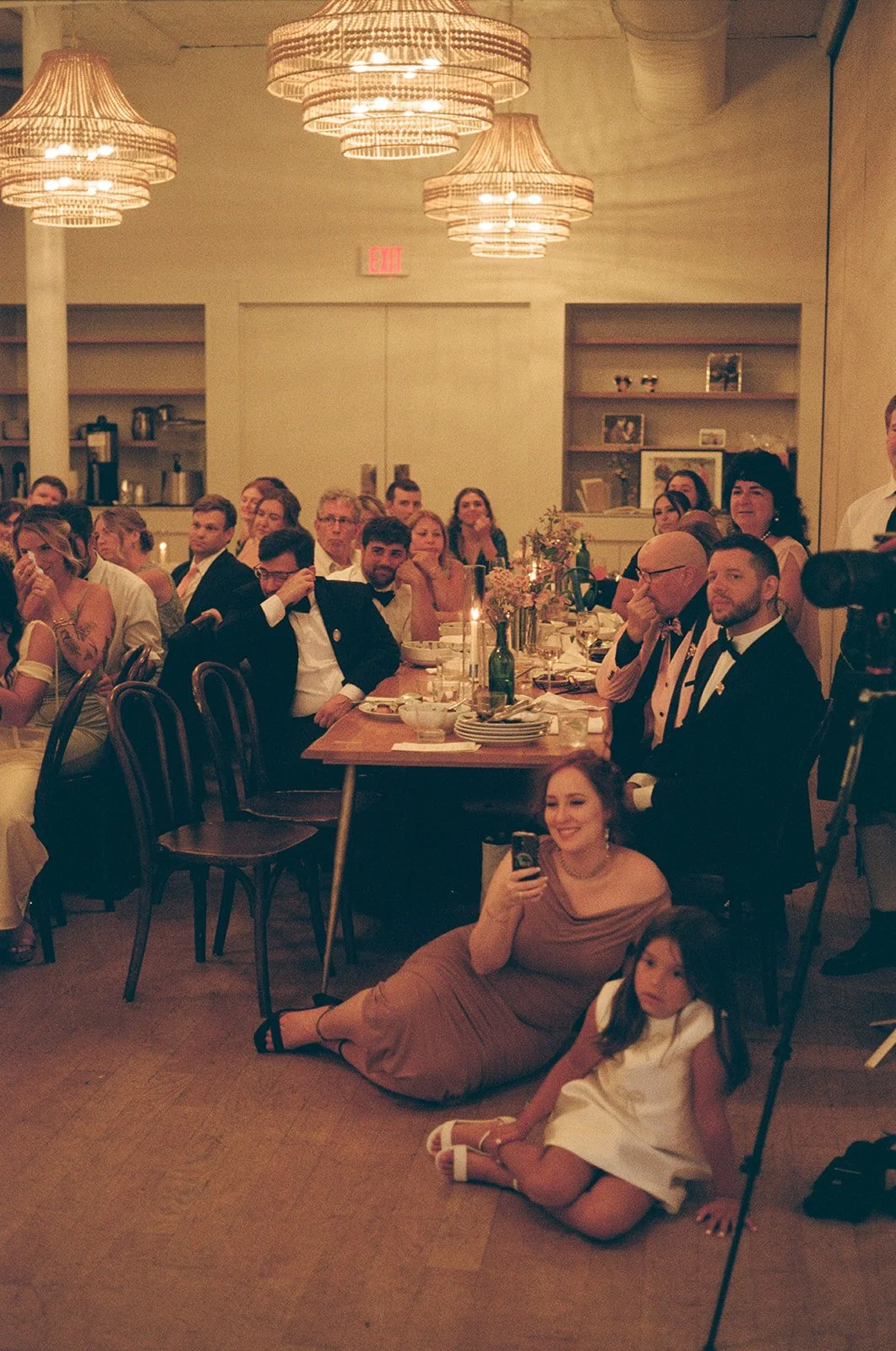 People dressed in formal attire attending a dinner party or celebration, with some sitting at a table and others standing, in a warmly lit room with modern chandeliers at Common House in Richmond Virginia.
