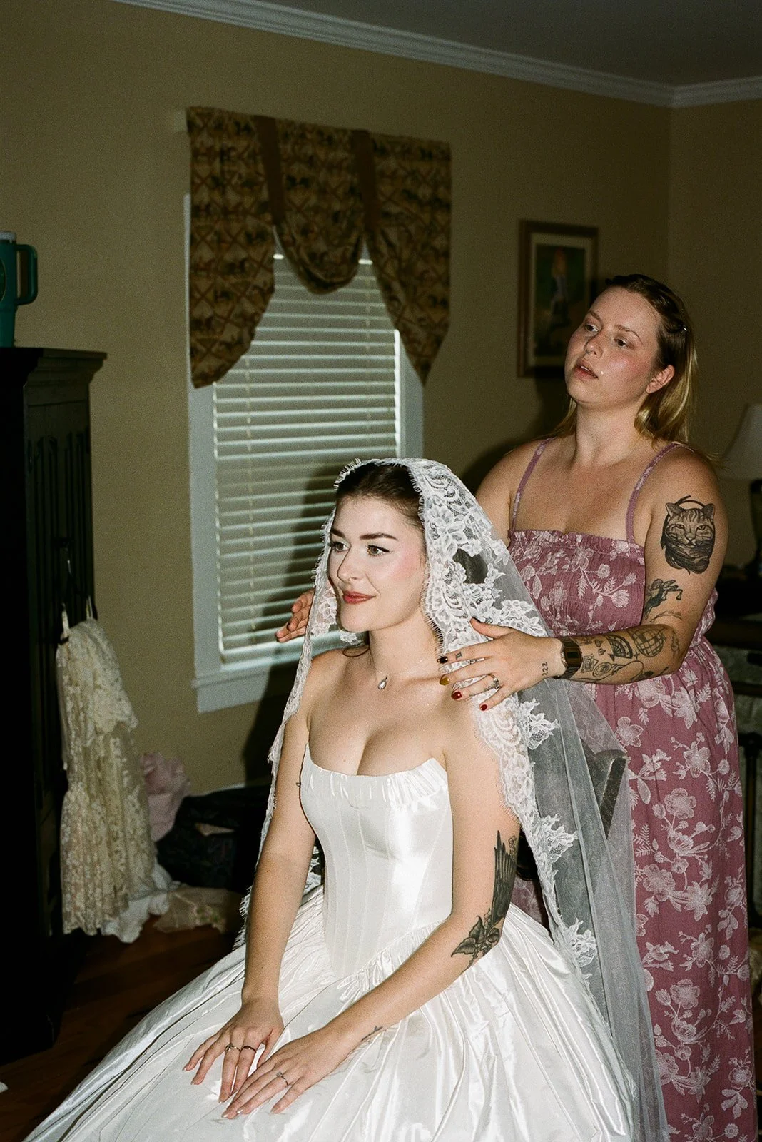 A bride sitting in a white wedding dress while a woman adjusts her lace veil in a room with beige walls and window blinds. charlottesville virginia wolftrap farm