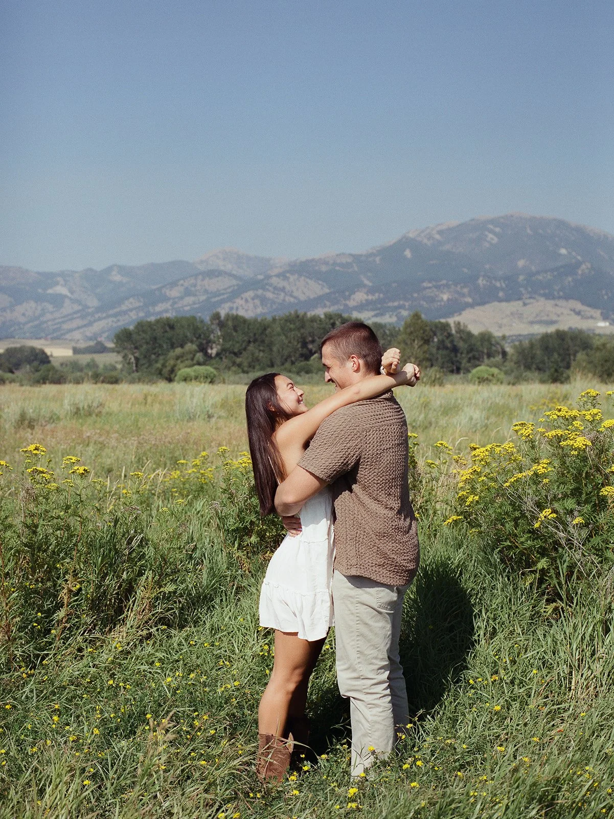 A couple embracing in a field of yellow flowers, with mountains in the background under a clear blue sky. bozeman montana engagement photographer film