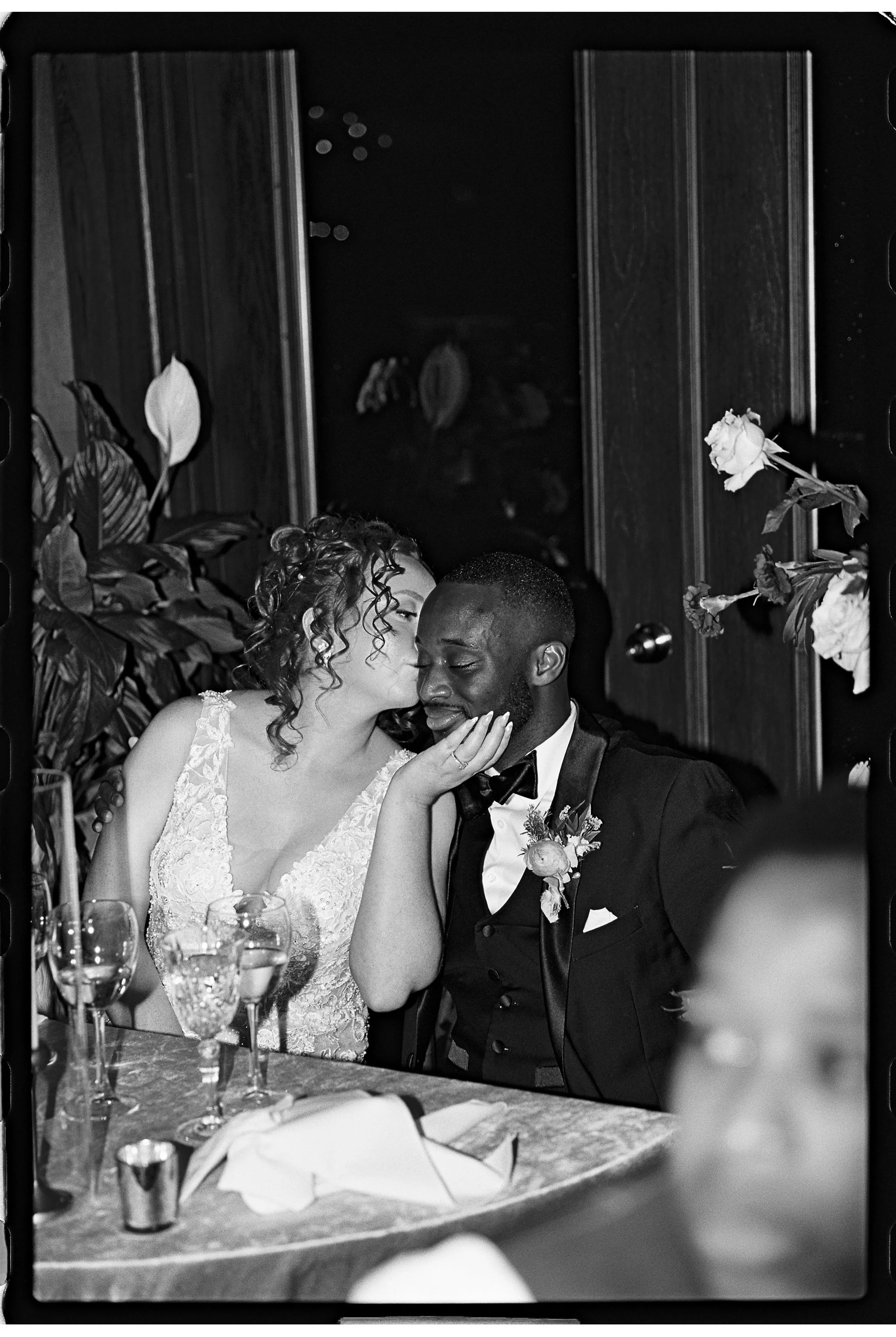 A black and white photo of a couple at a wedding, with the woman in a lacy wedding dress leaning in to kiss the man in a tuxedo. They are seated at a table with glasses and floral decorations.