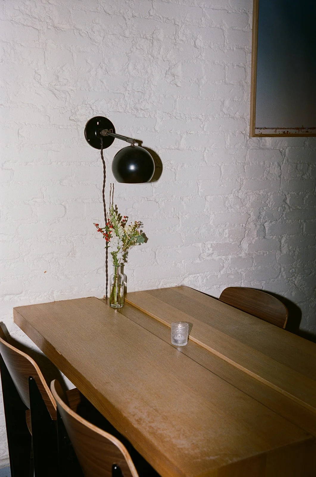 A wooden dining table with a glass vase containing flowers, a small candle holder, and a black wall-mounted lamp against a white brick wall. DC Wedding film photographer doubles cafe