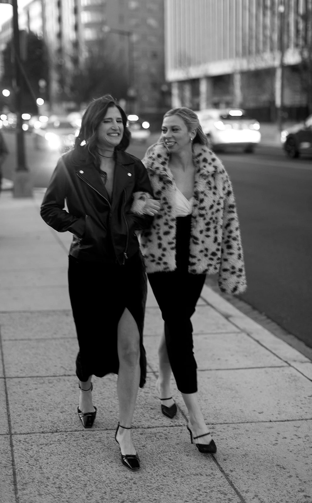 Two women walking on city sidewalk, one in a leather jacket and thigh-high slit skirt, the other in a leopard print fur coat and heels, smiling and enjoying each other's company. DC Wedding film photographer no kisses bar